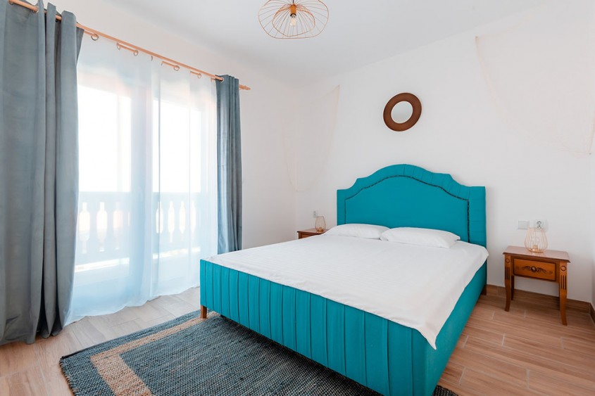 Dormitor în stil mediteranean cu pat turcoaz draperii vaporoase lumină naturală și mobilier din lemn decor